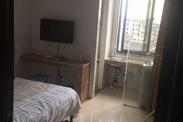 A bedroom with a bed, a desk, a chair, a TV, and a window with a view of buildings.