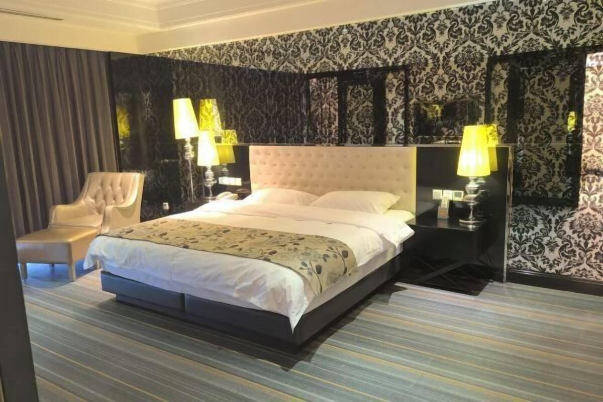 Una habitación de hotel con una cama grande, dos mesitas de noche con lámparas, una silla y un papel tapiz estampado.