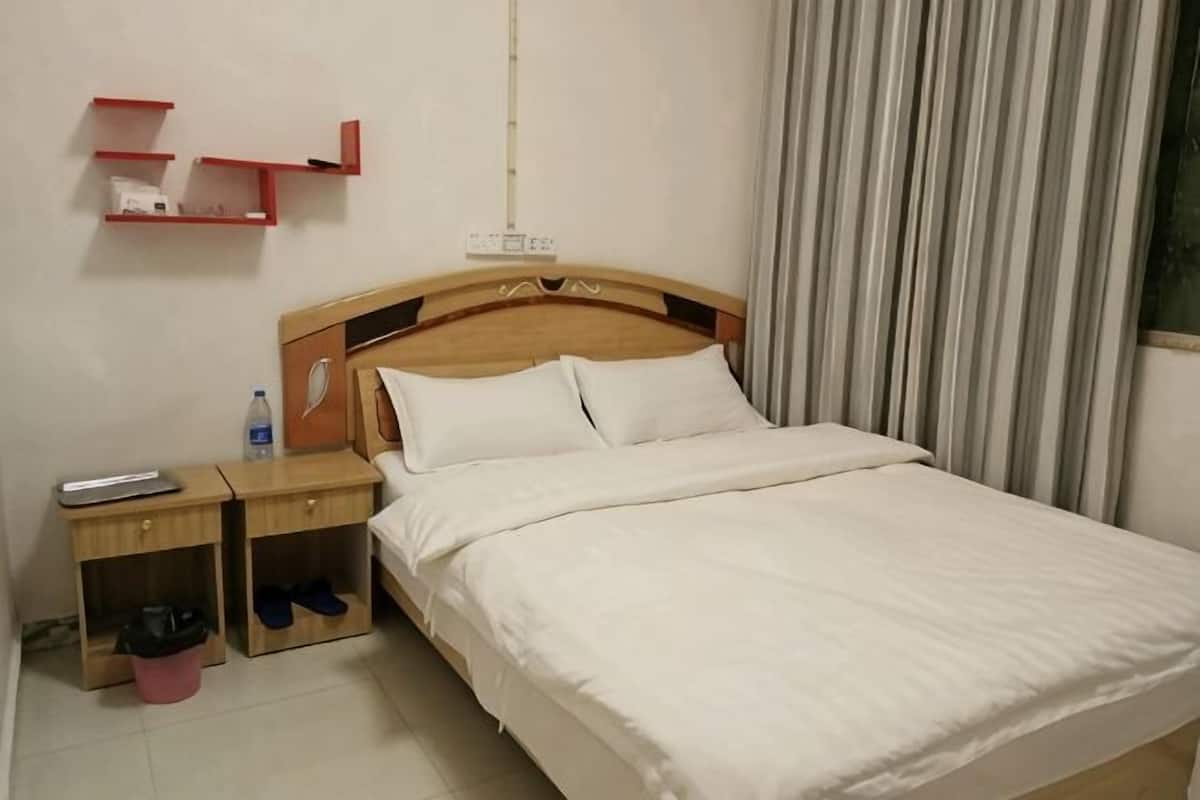 Ein Hotelzimmer mit einem Holz-Kopfteil, einem Einzelbett mit weißer Bettwäsche, einem Nachttisch mit Fernbedienung und einem roten Regal darüber.