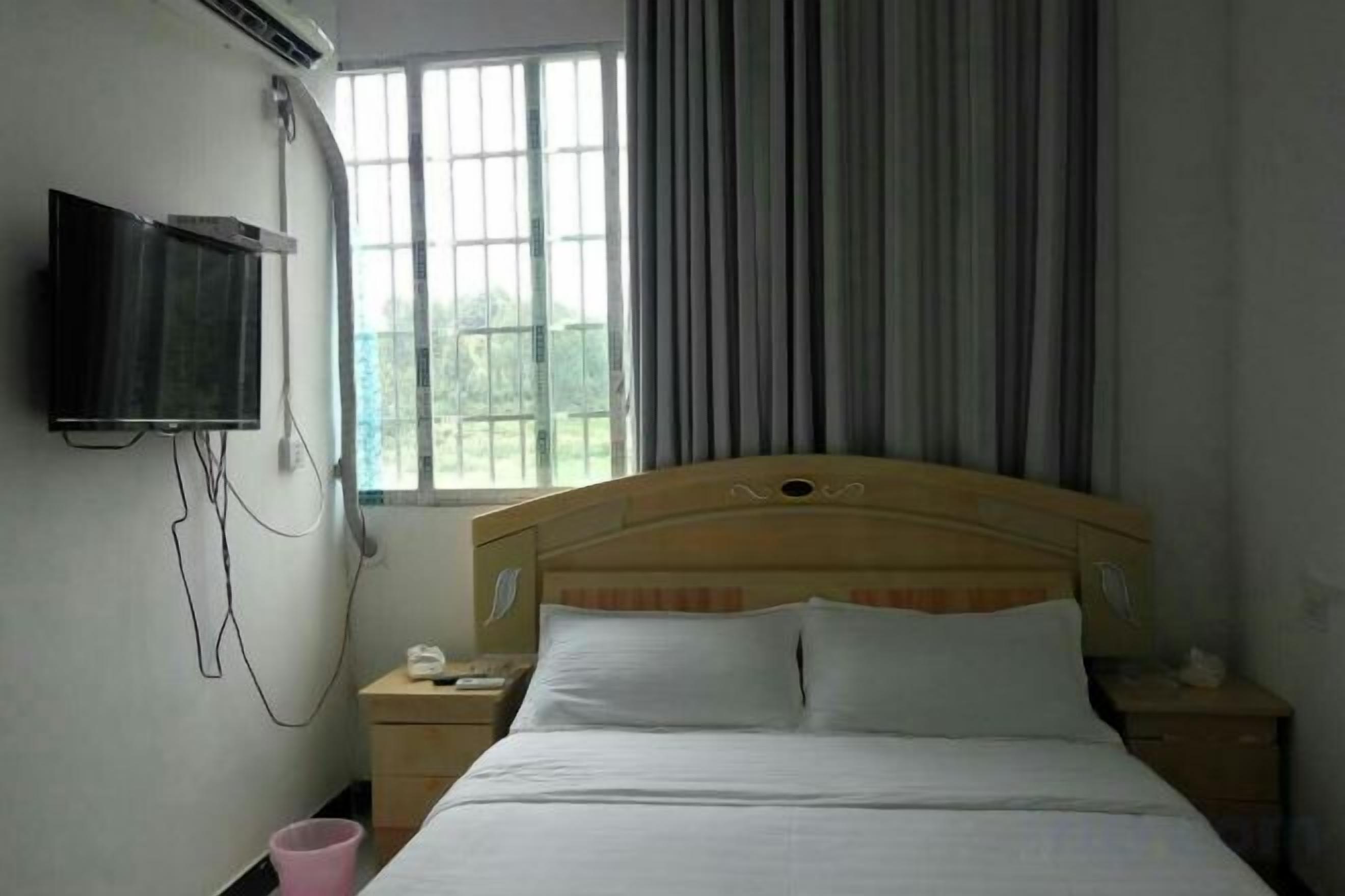 Une chambre d’hôtel avec un lit doté d’une tête de lit en bois, une table de chevet avec une lampe, un téléviseur fixé au mur et une fenêtre avec des rideaux.