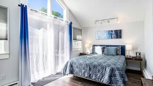 A bedroom with a large bed, a nightstand, a wall-mounted lamp, and a window with curtains. - Castaway Villas: Perfect Group Getaway – Heated Pool & Hot Tub Access (New Buffalo)