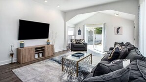 A modern living room with a large flat-screen TV, a wooden entertainment unit, a glass coffee table, a dark gray sofa, and a view of the outdoors through a glass door. - Castaway Villas: Perfect Group Getaway – Heated Pool & Hot Tub Access (New Buffalo)