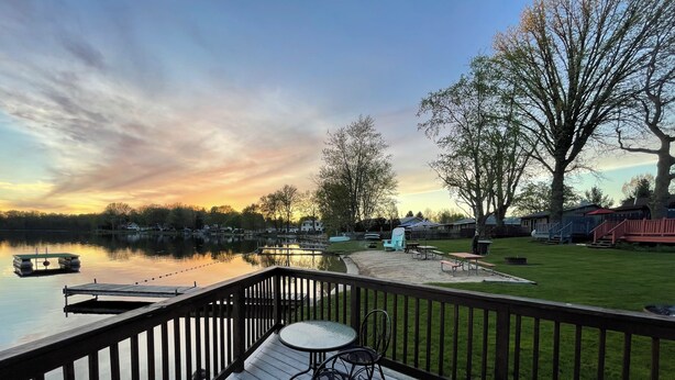 Una terraza de madera con una mesa y sillas, con vista a un lago tranquilo con un muelle y un área verde con mesas de picnic.