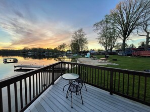 A wooden deck with a table and chairs, overlooking a calm lake with a dock and a grassy area with picnic tables.