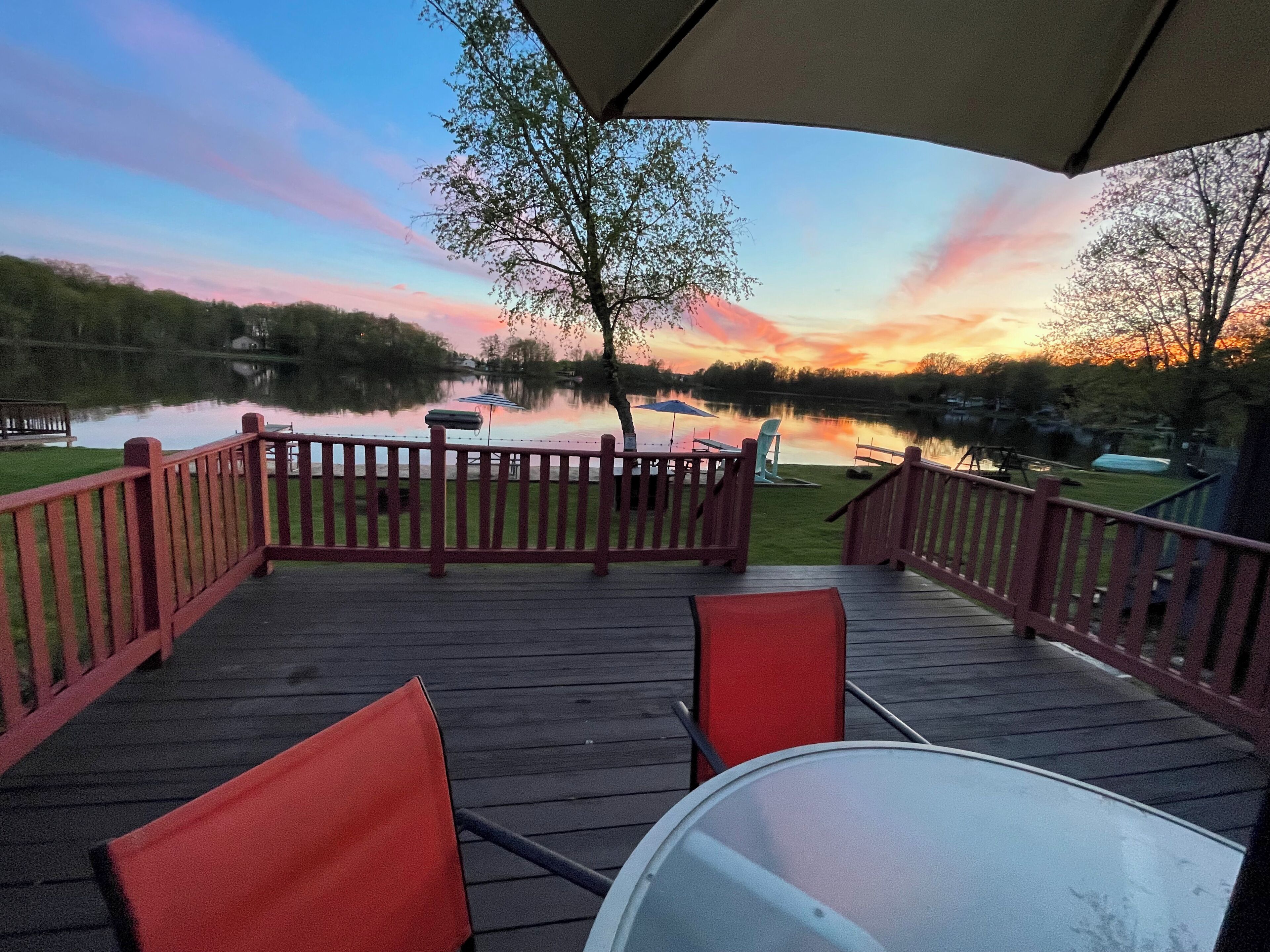 Terraza de madera con barandillas rojas, una mesa y sillas con vistas a un lago al atardecer.