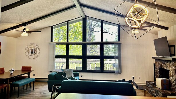 A spacious living area with a large window, a ceiling fan, a modern light fixture, and a stone fireplace.