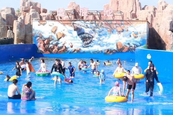 A water park with a large mural of a shark and waves, people in water, and inflatable rings.