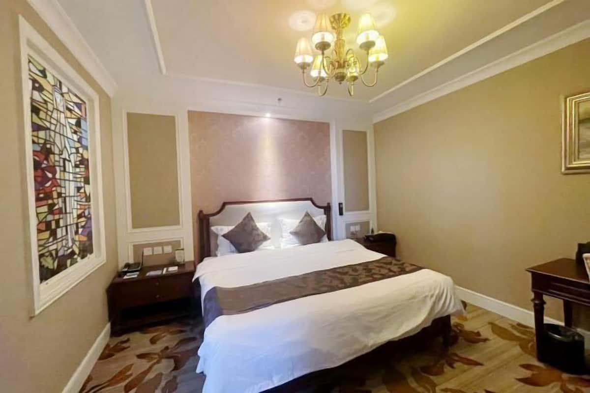Ein Hotelzimmer mit einem großen Bett, einem Nachttisch, einem Schreibtisch, einer Kristallleuchter und einem Buntglasfenster.