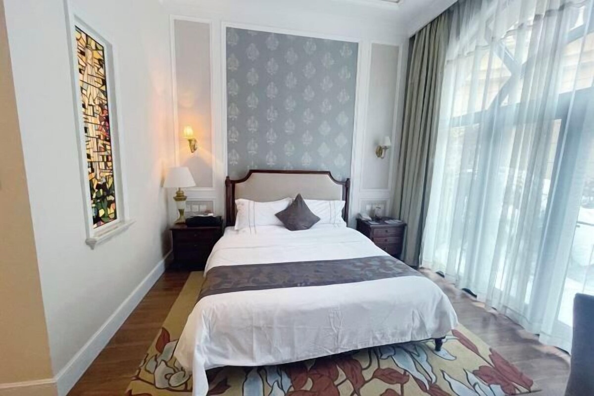 A hotel room with a large bed, a nightstand with a lamp, a wall-mounted light, a window with sheer curtains, and a patterned rug.