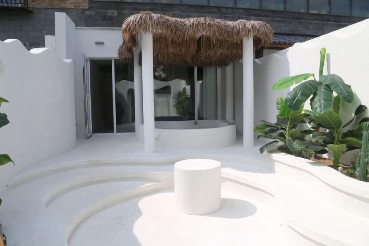 Un espace extérieur moderne doté d’une structure à toit de chaume, d’un espace de détente circulaire blanc et de plantes en pot.
