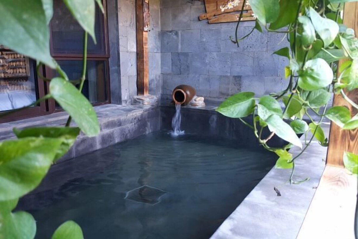 A stone-walled hot tub with a water spout, surrounded by greenery.