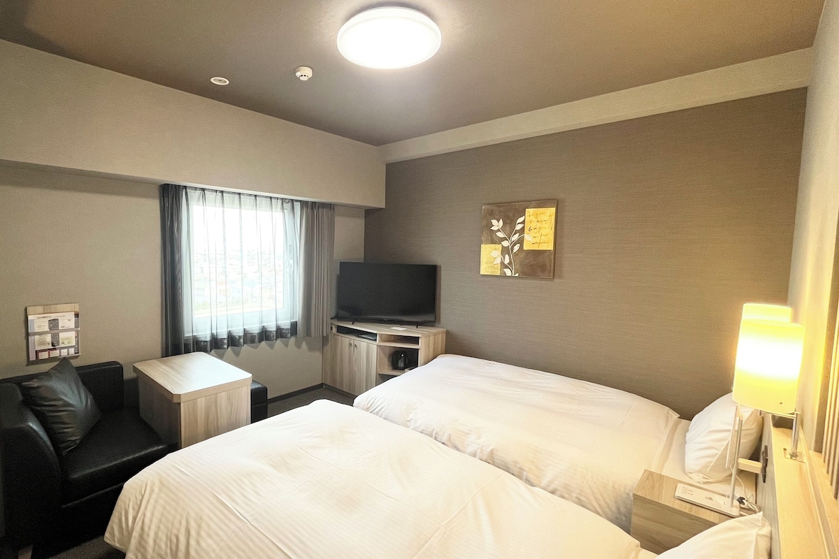 A hotel room with two beds, a desk with a flat-screen TV, a chair, and a lamp.