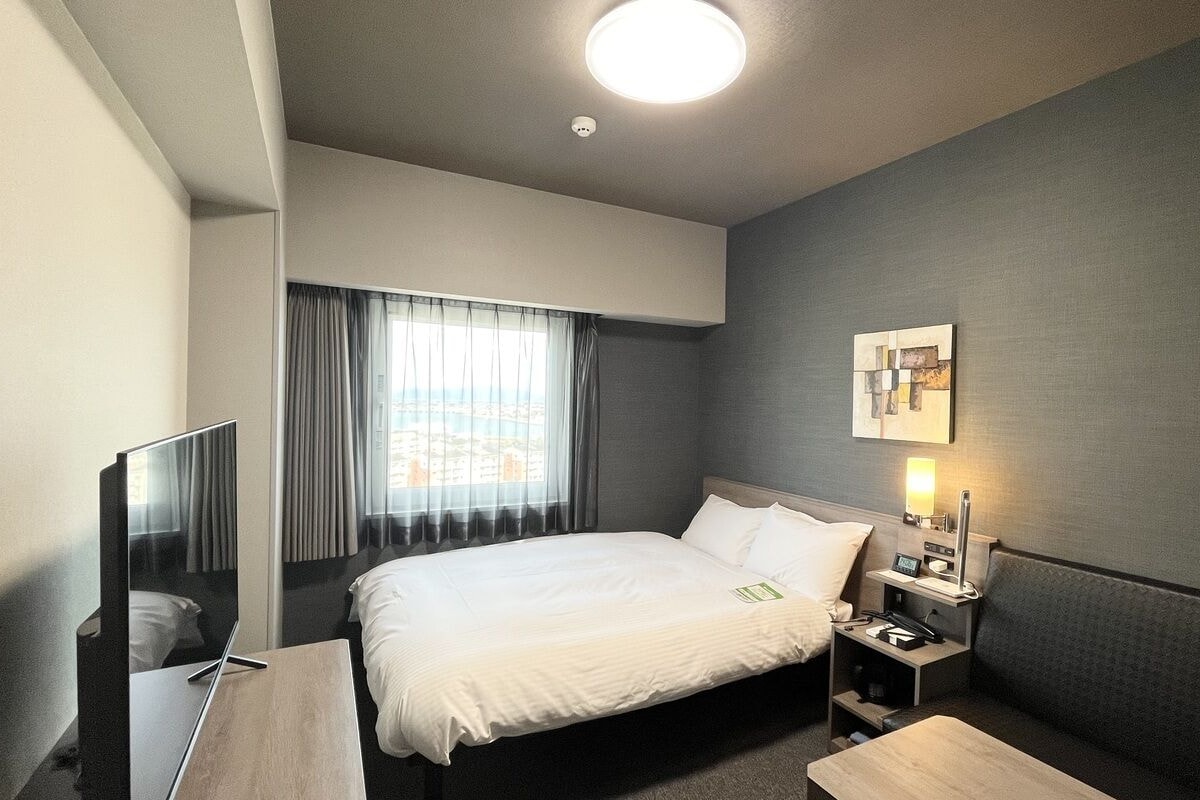 A hotel room with a bed, a desk, a chair, a TV, a bedside table with a lamp, and a window with curtains.