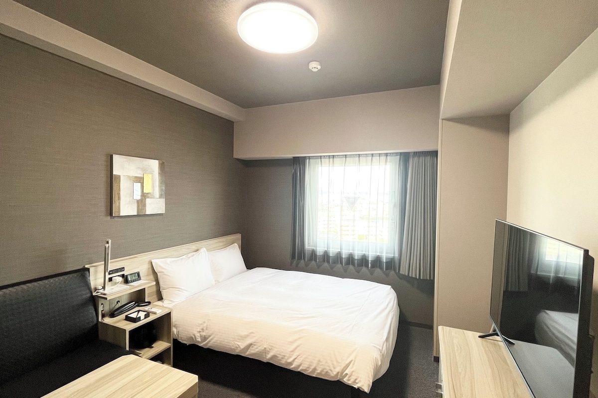 A hotel room with a bed, a bedside table, a TV, a window with curtains, and a ceiling light.