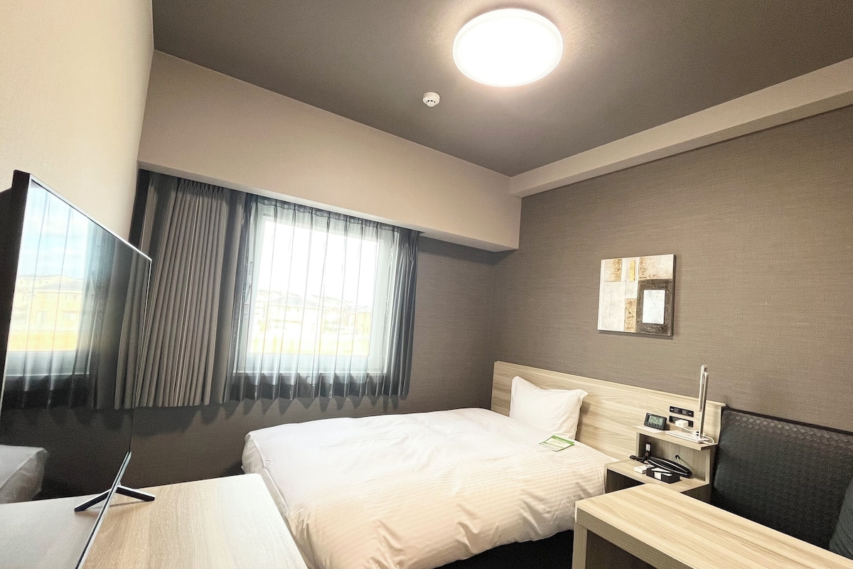 A hotel room with a bed, bedside table, TV, and a window with curtains.