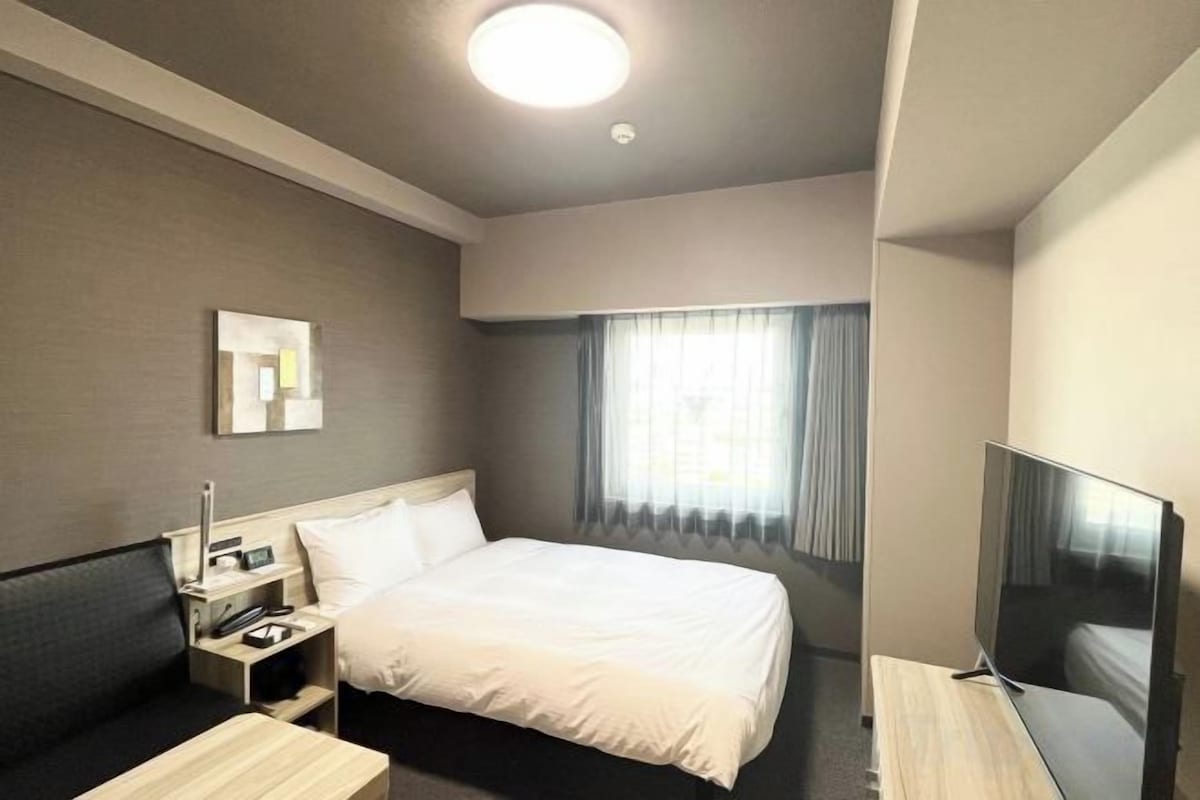 A hotel room with a bed, a TV, a desk, and a window with curtains.