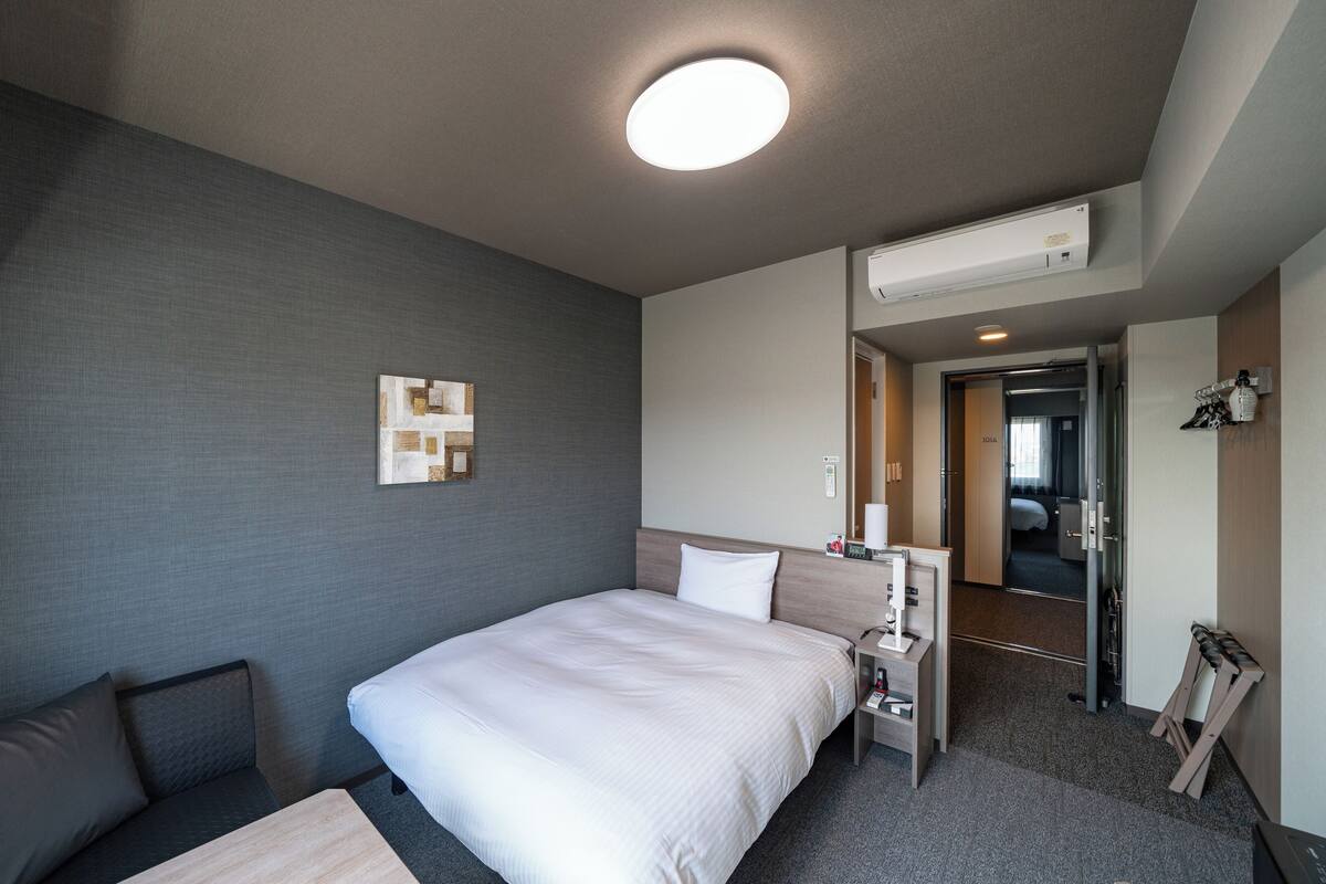 A hotel room with a bed, a desk, a chair, a nightstand, a mirror, and a door leading to another room.