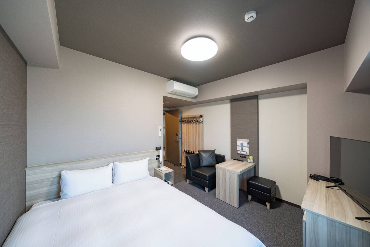 A hotel room with a large bed, a desk, a chair, a TV, and an air conditioning unit.