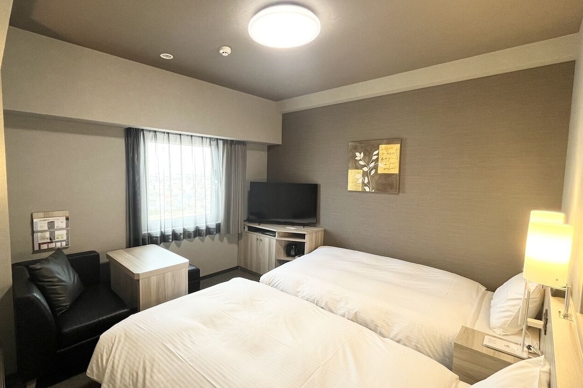 A hotel room with two beds, a desk, a chair, a television, and a lamp.