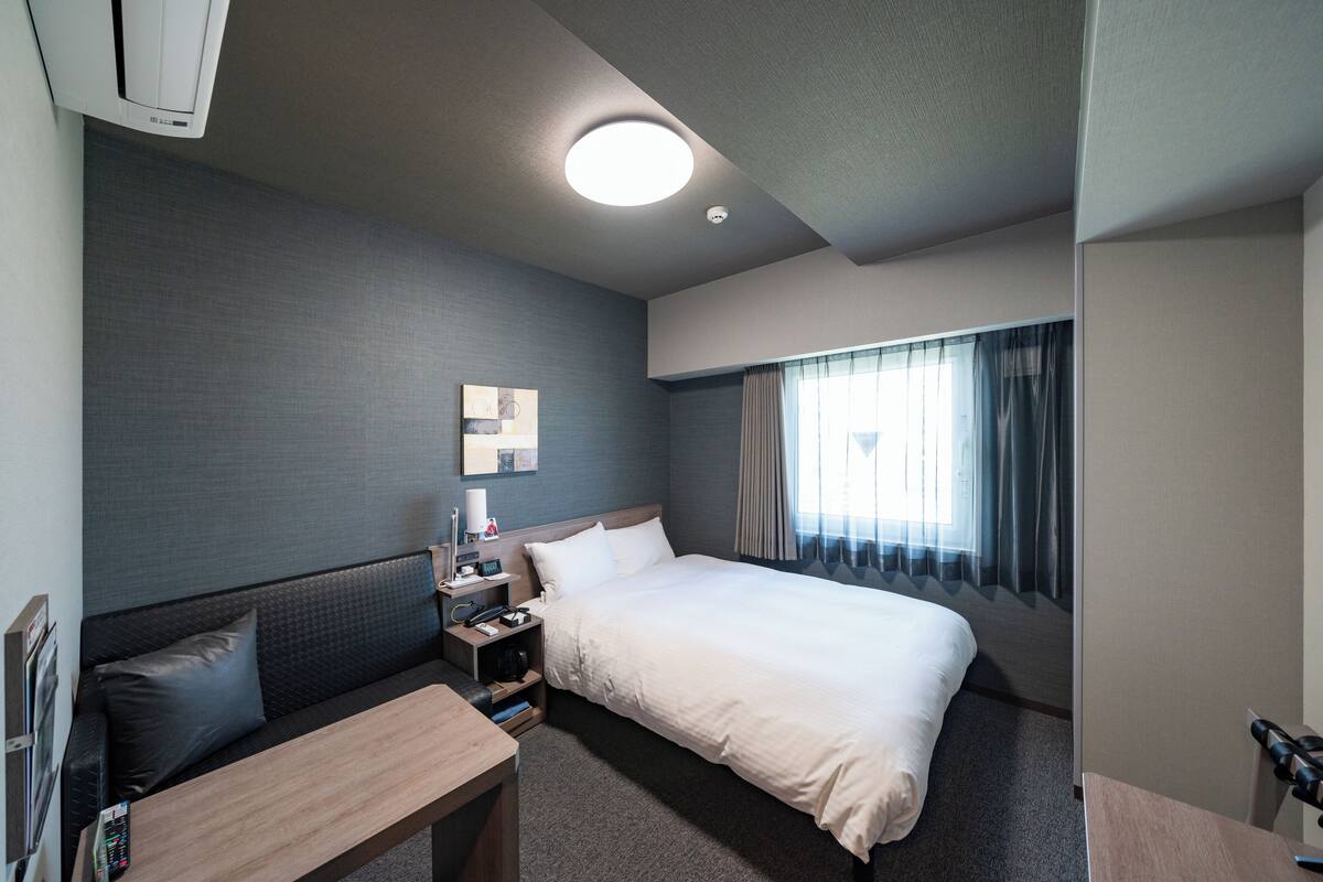 A hotel room with a bed, a bedside table, a desk, a chair, a sofa, a TV, and a window with curtains.
