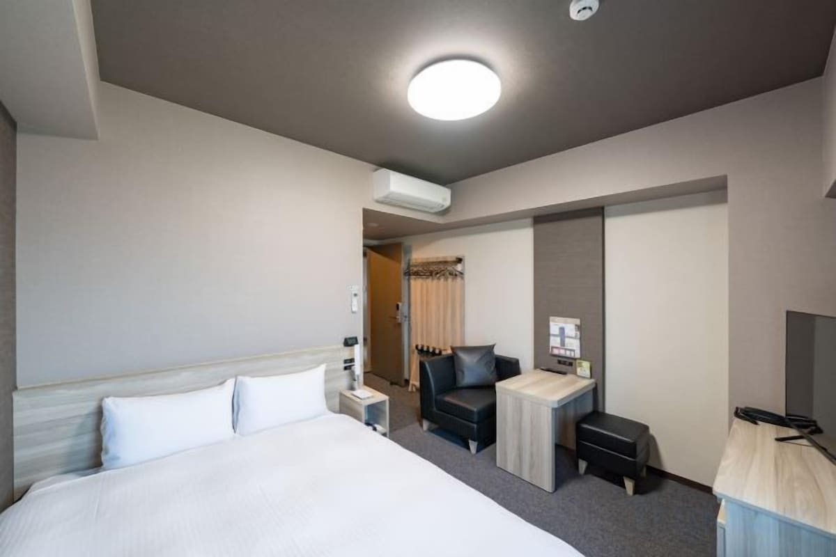 A hotel room with a bed, a desk, a chair, a TV, and an air conditioning unit.