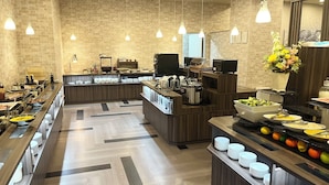 Ein Frühstücksbereich mit Buffet und mehreren Servicestationen, einer Ziegelwand im Hintergrund und Pendelleuchten.