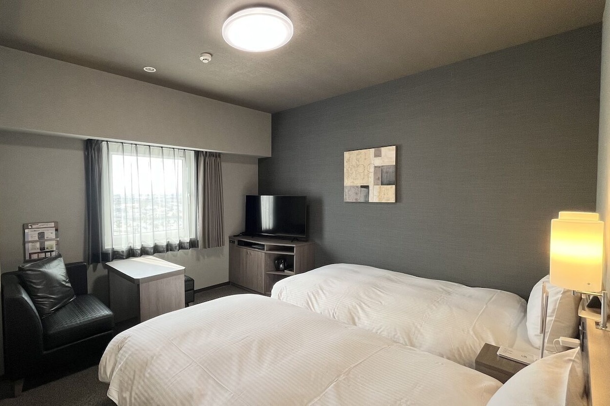 A hotel room with two beds, a desk, a chair, a TV, and a lamp.