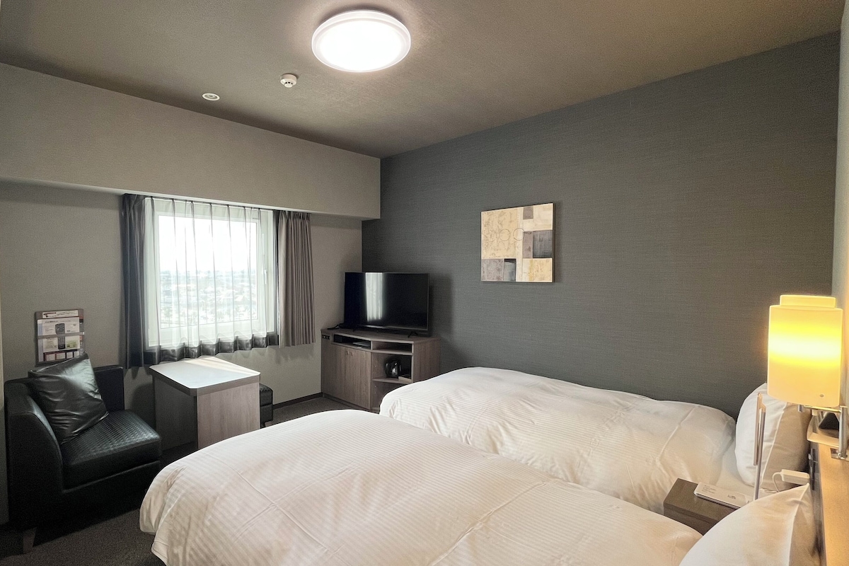 A hotel room with two beds, a desk, a chair, a television, and a window with curtains.
