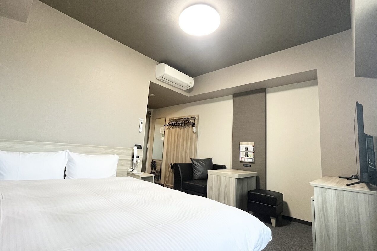 A hotel room with a large bed, a desk, a chair, an air conditioning unit, and a television.