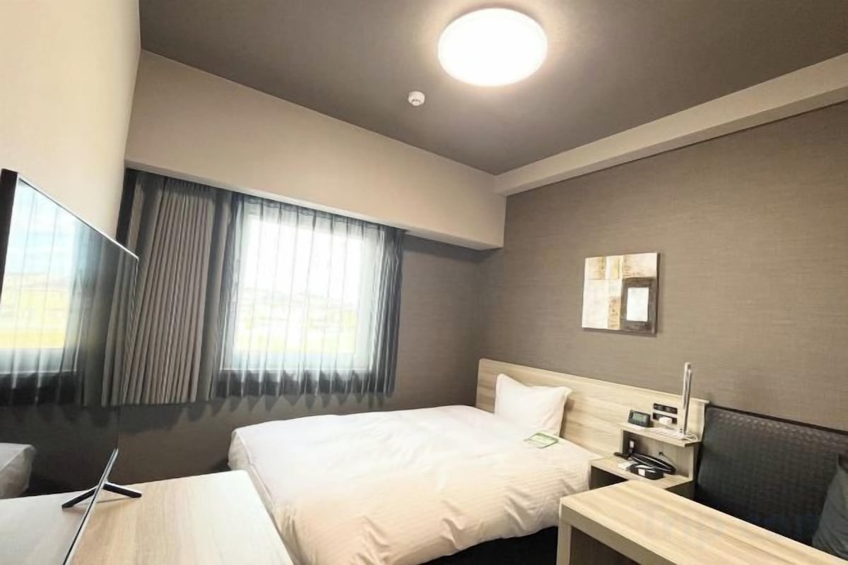 A hotel room with two beds, a desk, a chair, a TV, and a window with curtains.