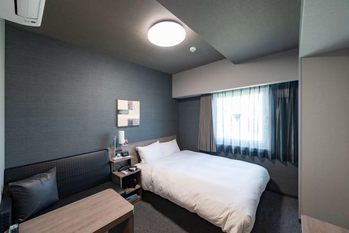 A hotel room with a bed, bedside table, lamp, and a window with curtains.