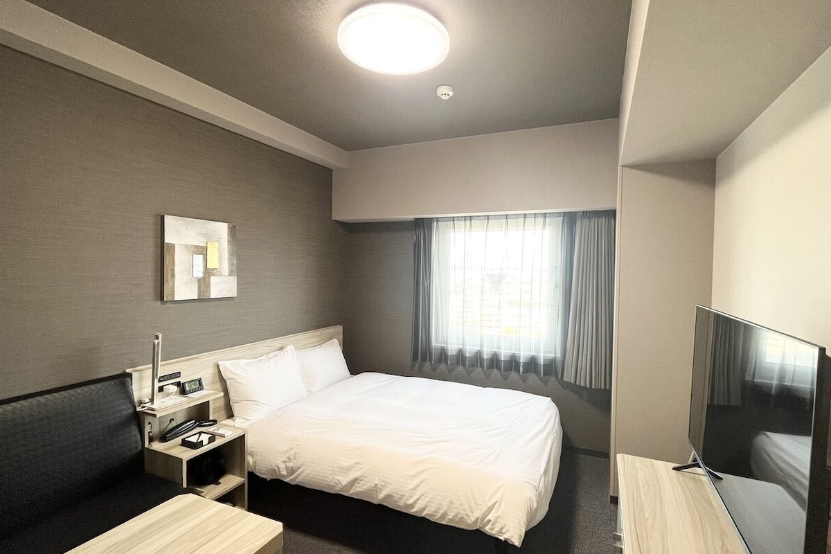 A hotel room with a bed, bedside table, TV, and a window with curtains.