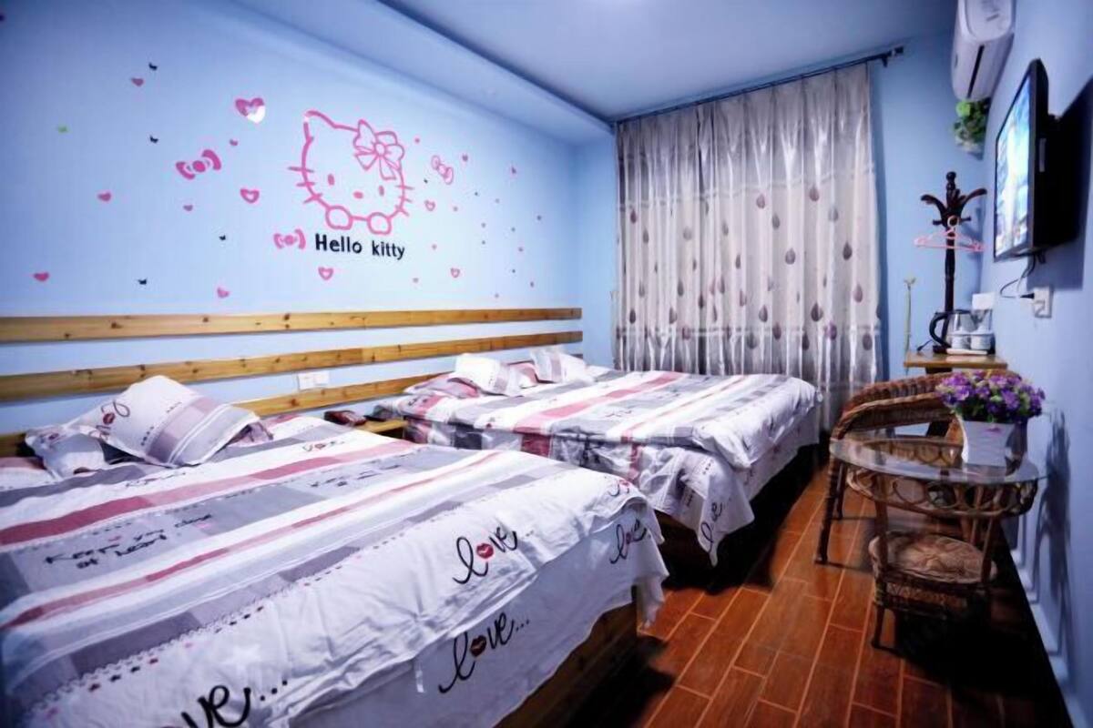 A hotel room with two beds, a wall decal of Hello Kitty, a window with curtains, and a small table with a vase.