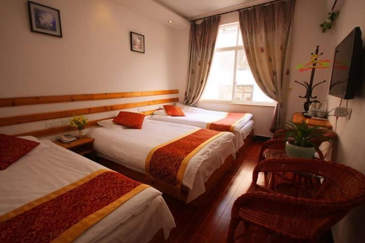 A hotel room with three single beds, a wooden headboard, a small table with a plant, and a wicker chair.