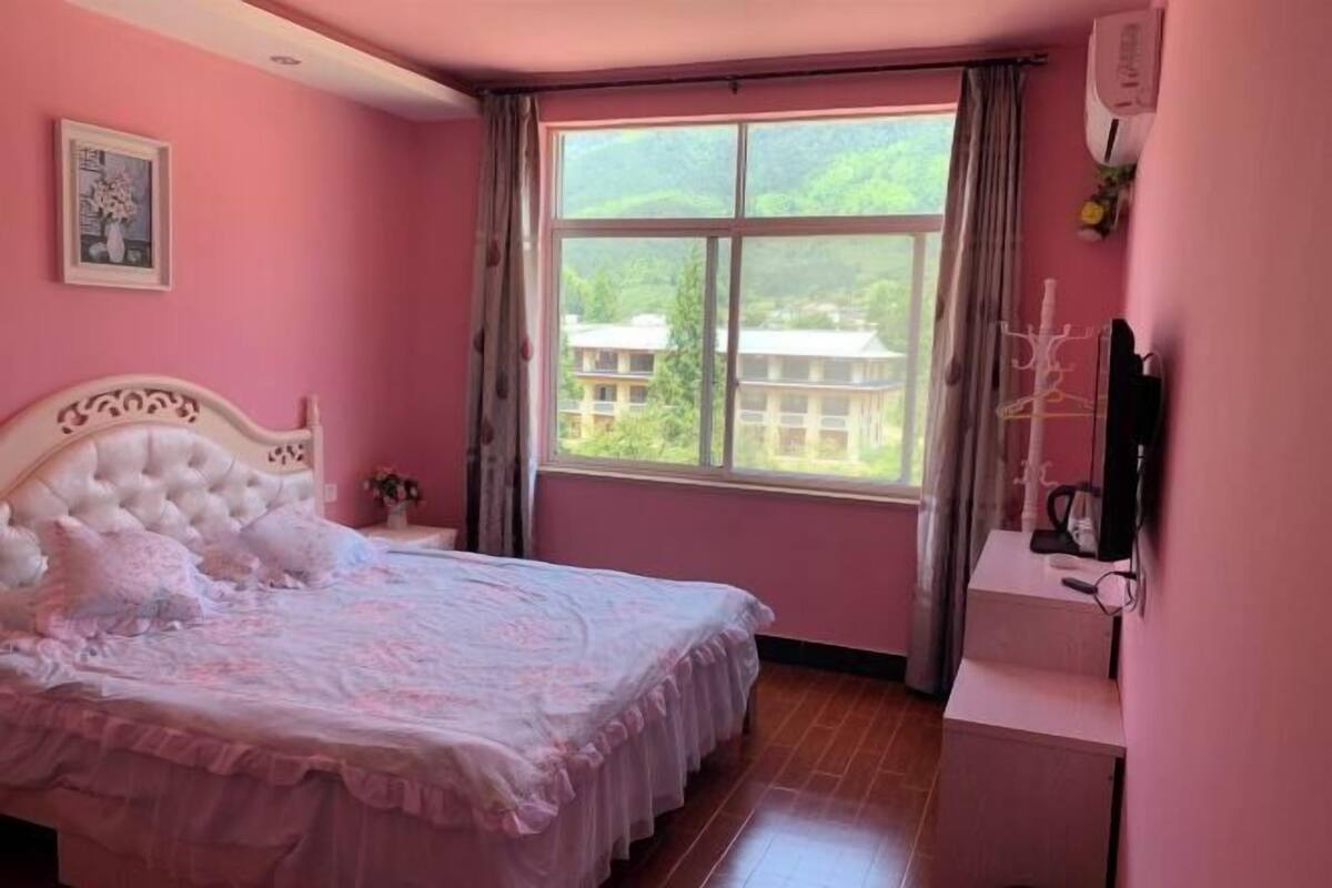 A bedroom with a large bed, a window with a view of buildings, a bedside table, and a wall-mounted TV.