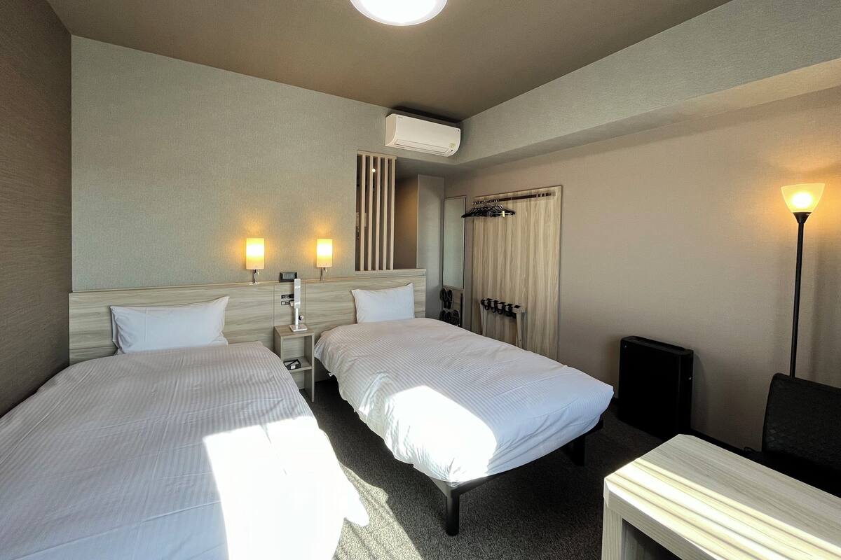 Ein Hotelzimmer mit zwei Einzelbetten, einem Nachttisch, einem Schreibtisch, einem Stuhl, einer Lampe und einer Klimaanlage.