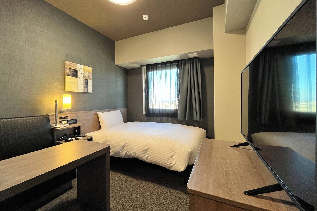 Ein Hotelzimmer mit einem Bett, einem Schreibtisch, einem Fernseher, einer Lampe und einem Fenster mit Vorhängen.