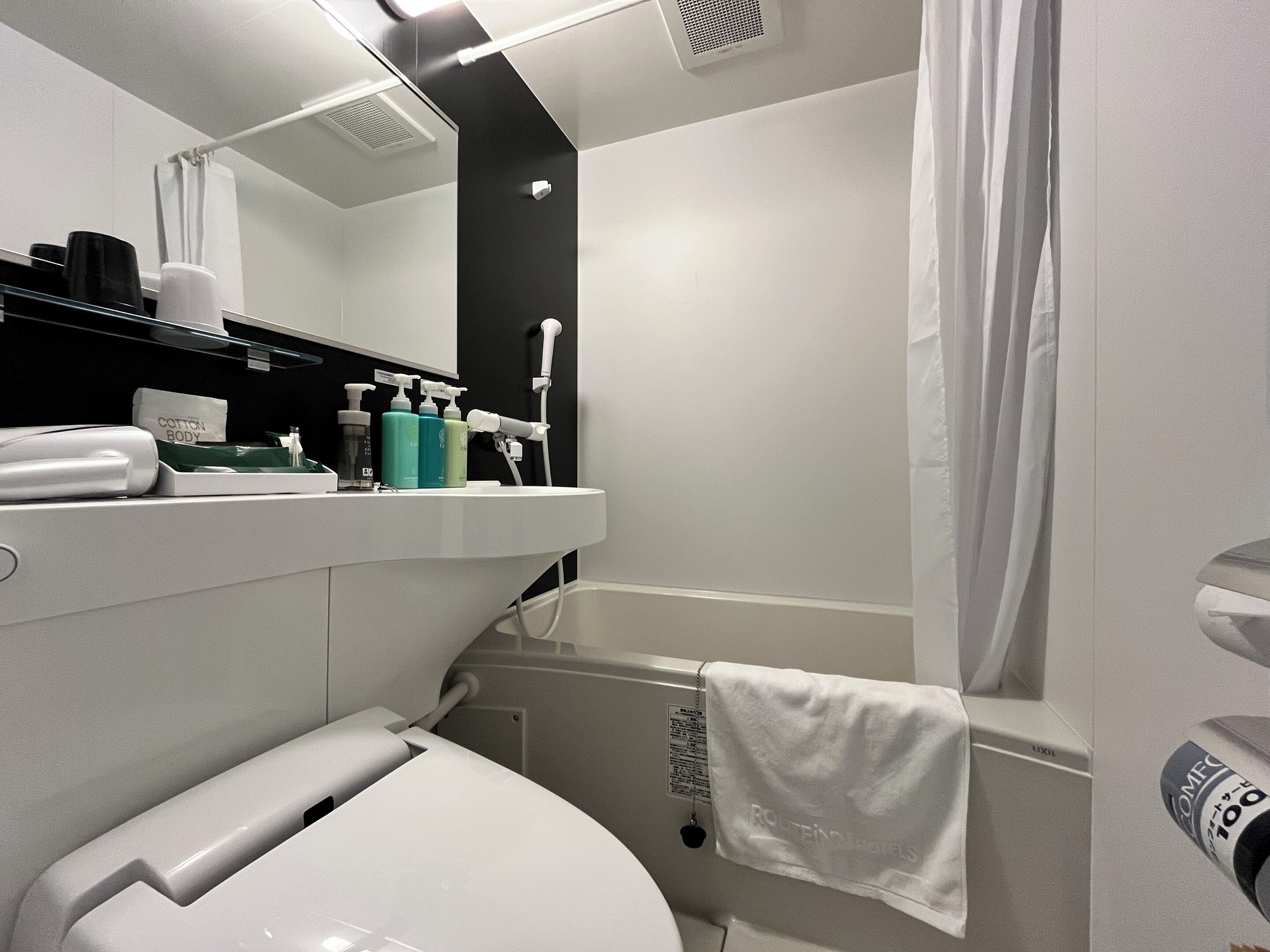 A modern bathroom with a toilet, sink, and shower area. There are toiletries on the sink and a towel hanging on the shower curtain.