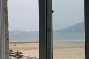 Vista a la playa desde una ventana, que incluye una playa de arena, agua tranquila y montañas a lo lejos.
