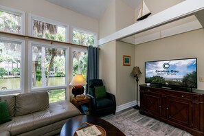 A living room with a sofa, a television, a lamp, and a view of a water body through large windows.