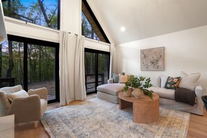 A modern living room with a large window, a sofa, a coffee table, and a view of a forest. - Uckor's Cottage | Hocking Hills, 5 Acres, Hot Tub (Rockbridge)