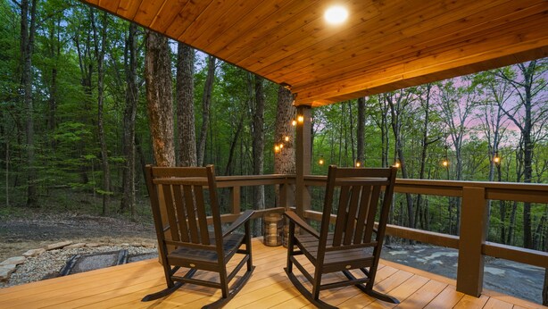 Eine Holzterrasse mit zwei Schaukelstühlen, einem Tisch und einem Geländer mit Blick auf einen Wald.
