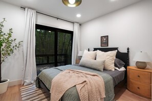 A bedroom with a large window, a bed with pillows, a bedside table with a lamp, and a potted plant. - Uckor's Cottage | Hocking Hills, 5 Acres, Hot Tub (Rockbridge)