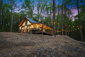 A cabin with a stone exterior, wooden deck, and string lights. - Uckor's Cottage | Hocking Hills, 5 Acres, Hot Tub (Rockbridge)
