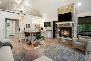 A modern living room with a stone fireplace, a kitchen area, and a dining space. - Uckor's Cottage | Hocking Hills, 5 Acres, Hot Tub (Rockbridge)