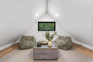 A cozy attic room with a sloped ceiling, a large window offering a forest view, two green bean bags, a coffee table with a vase and a tray, and a patterned rug. - Uckor's Cottage | Hocking Hills, 5 Acres, Hot Tub (Rockbridge)