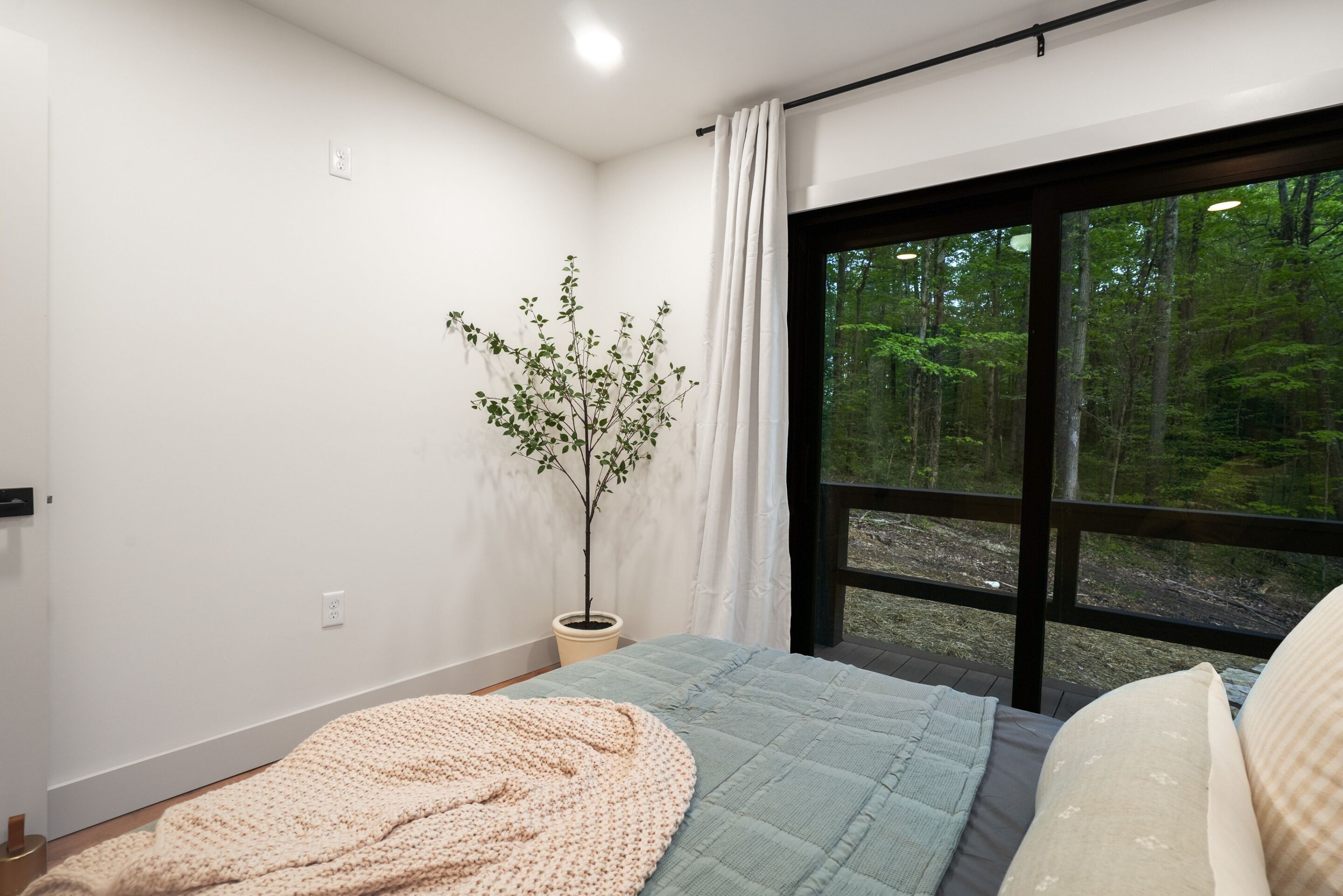 Une chambre avec une grande fenêtre, une plante en pot et un lit recouvert d’une couverture bleue et beige.