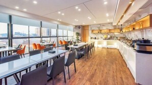A modern dining area with long tables and chairs, a kitchen area, and large windows offering a city view.