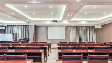 Un salón de conferencias con filas de mesas y sillas, una pantalla de proyector y una pantalla de presentación.