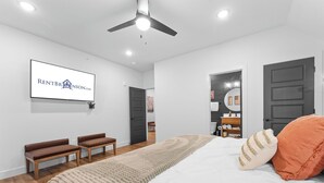 Camera da letto moderna con un letto, due panche, una ventola a soffitto e una TV a muro che mostra il logo di un'agenzia di affitti.